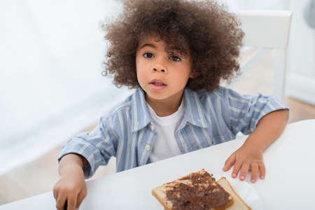 Toddler african american kid looking at camera near bread with chocolate paste in kitchenの写真素材