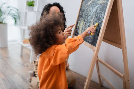 Toddler african american girl drawing on chalkboard near mom at homeの写真素材