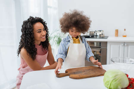 Positive african american mother in apron standing near daughter, chopping board and fresh vegetables at homeの写真素材