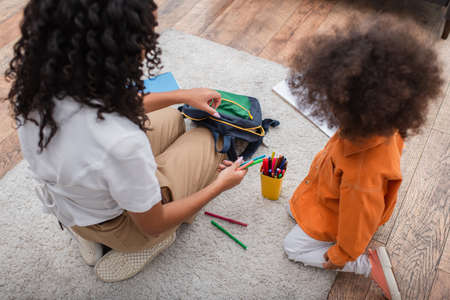 Overhead view of african american mom holding color pencils and backpack near kid at homeの写真素材