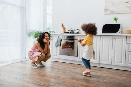 Smiling african american mom looking at daughter in apron in kitchenの写真素材