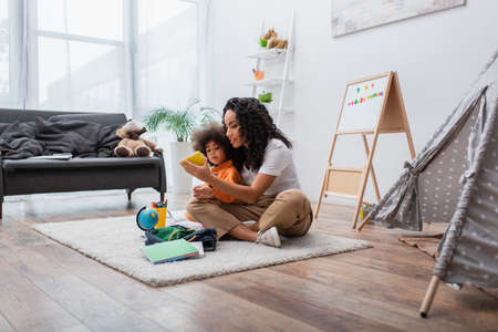 African american mom holding notebook near kid, stationery and globe on carpet at homeの写真素材