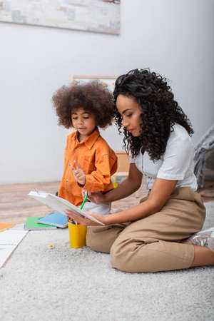 African american mom holding book near toddler kid and notebook at homeの写真素材