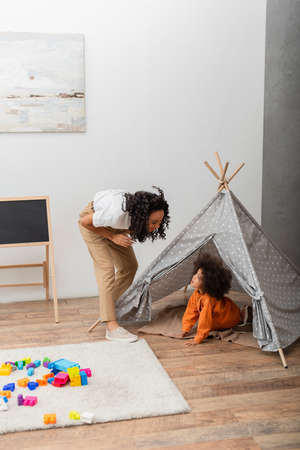 African american mom looking at toddler kid in teepee at home