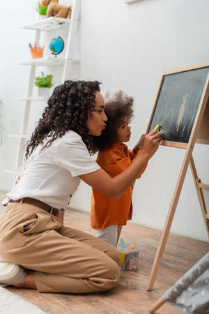 Young african american woman drawing on chalkboard with daughter at homeの写真素材