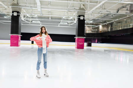full length of happy woman in white scarf and ear muffs showing heart with hands while skating on...