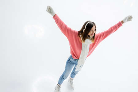 high angle view of of excited young woman in ear muffs and jeans skating with outstretched hands on...