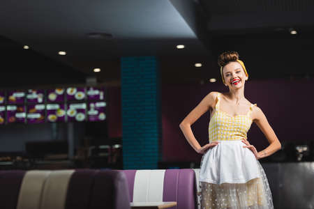 happy pin up waitress standing with hands on hips in cafe