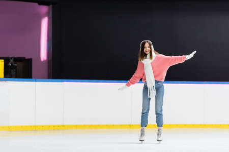full length of smiling woman in ear muffs and winter outfit skating on ice rinkの写真素材