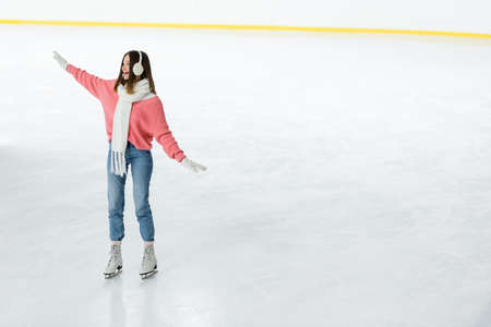 full length of amazed young woman in ear muffs and scarf skating with outstretched hands on ice rinkの写真素材