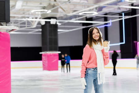 happy woman in ear muffs and pink sweater holding paper cup on ice rinkの写真素材