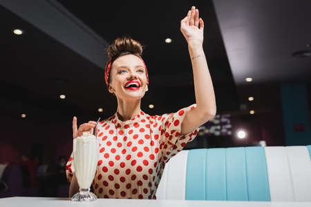 positive pin up woman in red polka dot blouse gesturing near milkshake on table