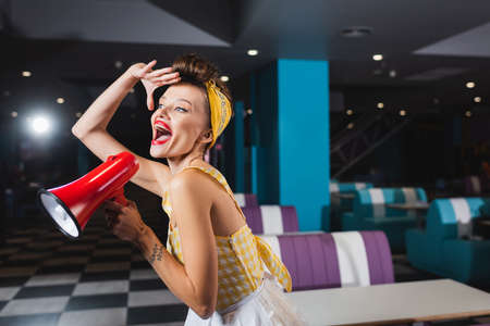 amazed pin up woman screaming in megaphone in retro cafe