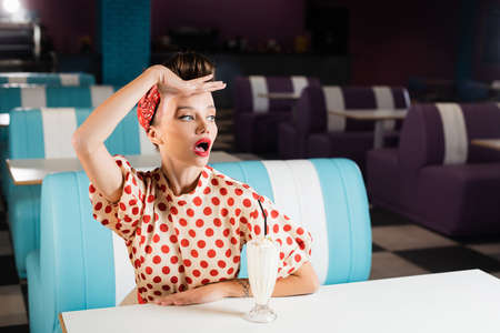 surprised pin up woman looking away near milkshake on tableの写真素材