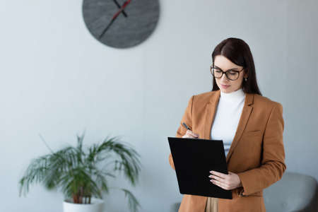 psychologist in eyeglasses writing on clipboard near blurred plant and wall clock