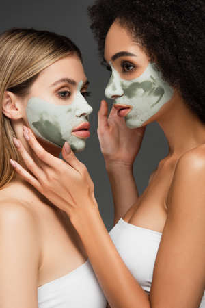multiethnic women in facial masks touching faces of each other isolated on greyの写真素材