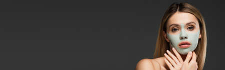 young woman looking at camera while pampering face with clay mask isolated on grey, bannerの写真素材