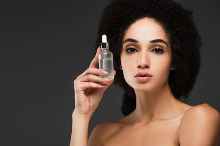 pretty african american woman with bare shoulders holding bottle of cosmetic serum isolated on greyの写真素材