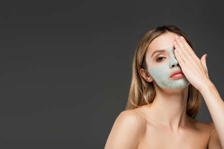 young woman with naked shoulders and clay mask obscuring face with hand isolated on greyの写真素材