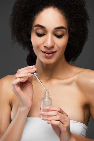 smiling african american woman with clean skin and natural makeup holding cosmetic serum isolated on greyの写真素材