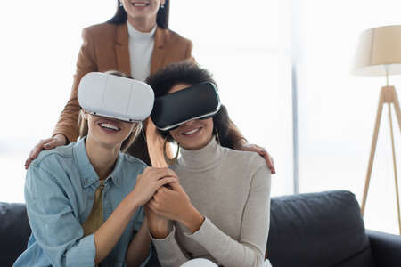 smiling interracial lesbian couple in vr headsets holding hands near blurred psychologist