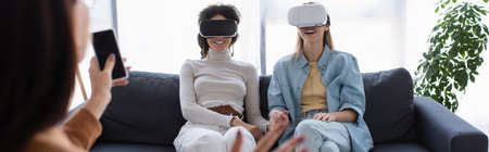 interracial lesbian couple in vr headsets smiling near blurred psychologist with smartphone, bannerの写真素材