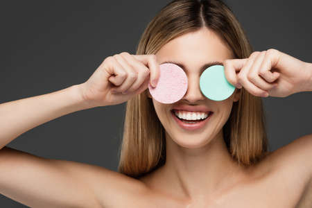 young woman smiling while covering eyes with cosmetic sponges isolated on greyの写真素材