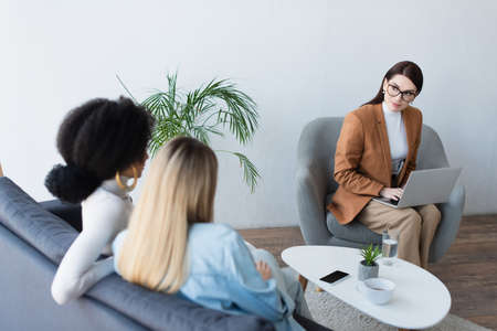 psychologist typing on laptop while looking at interracial lesbian couple sitting on sofaの写真素材