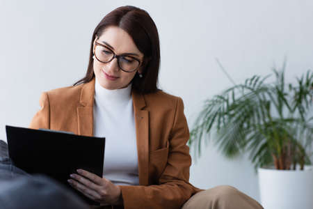 smiling psychologist in eyeglasses sitting with clipboard in consulting room