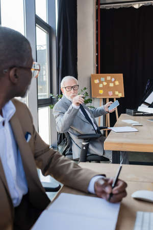 elderly businessman with cellphone talking to african american colleague working on blurred foregroundの写真素材
