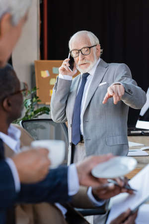 elderly businessman pointing with finger while talking on mobile phone near blurred colleaguesの写真素材