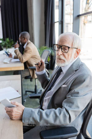elderly businessman in eyeglasses holding mobile phone while african american colleague working on blurred backgroundの写真素材