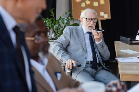 senior businessman sending voice message on smartphone near blurred multiethnic colleagues