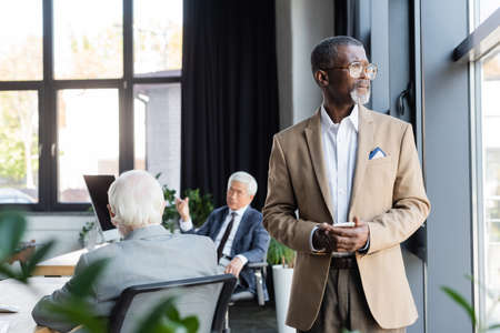 african american businessman with smartphone standing near window while blurred colleagues working in officeの写真素材