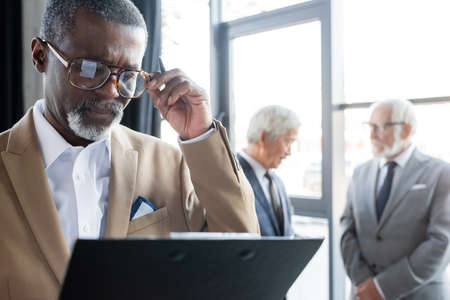 senior african american businessman with clipboard touching eyeglasses while colleagues talking on blurred backgroundの写真素材