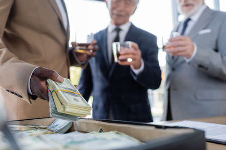 cropped view of african american businessman holding dollars near briefcase and blurred senior colleagues with glasses of whiskeyの写真素材