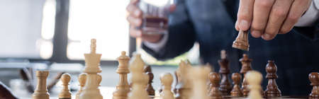 cropped view of businessman with blurred glass of whiskey playing chess in office, bannerの写真素材