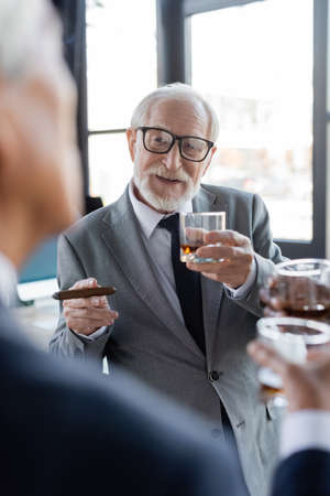 senior businessman with cigar and glass of whiskey talking to blurred colleague in officeの写真素材