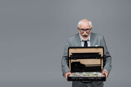astonished senior businessman looking at camera while holding briefcase with money isolated on grey