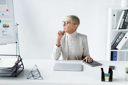 pensive senior businesswoman looking away near gadgets on deskの写真素材