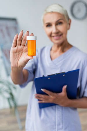 smiling and blurred nurse holding bottle with pills and clipboardの写真素材