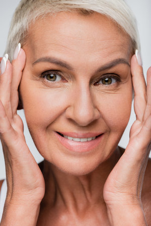 portrait of senior woman touching face while smiling isolated on greyの写真素材