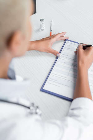 high angle view of blurred doctor writing prescription near syringe on desk