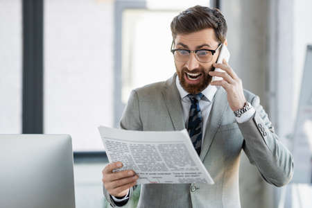 Excited businessman talking on smartphone while reading news in officeの写真素材