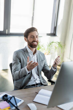 Positive businessman pointing with hand during video call on computer near notebook in office
