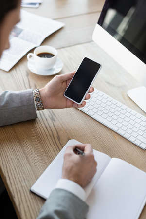 Businessman holding cellphone with blank screen and writing on notebook near computer in officeの写真素材