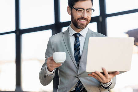 Cheerful businessman holding cup and laptop in officeの写真素材