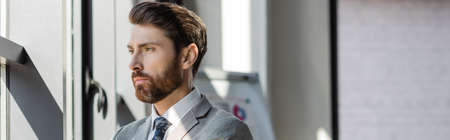 Bearded businessman in suit standing near window in office, banner