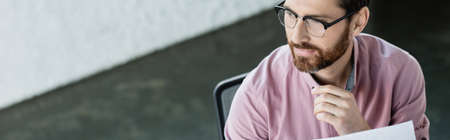 Bearded businessman in eyeglasses looking away near paper in office, banner