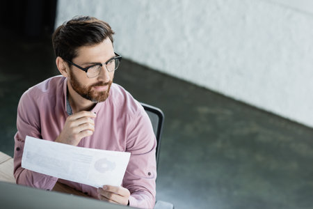 High angle view of businessman in shirt holding document near computer monitor in officeの写真素材